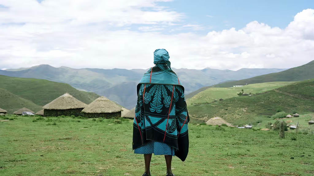 Indivíduo com trajes tradicionais basotho de virado costas, observando a paisagem vasta de um vilarejo nas montanhas Maluti. Imagem finalista do Grand Prix de Fotografia do Festival de Inverno de Hyères em 2002, criada por Joël Tettamanti.