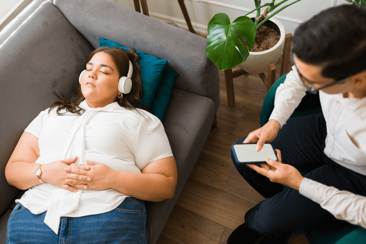 Ângulo alto de uma mulher deitada no sofá do terapeuta, ouvindo com fones de ouvido uma meditação guiada para relaxar com exercícios respiratórios.