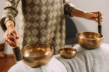 Mulher tocando tigelas tibetanas, posicionadas em cima das costas de uma pessoa deitada, em sessão terapêutica de sound healing.